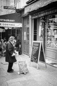An older woman with a shopping cart stands in front of a deli store, reading a chalkboard menu on the sidewalk; nearby, people sit at outdoor tables. The black-and-white scene captures an everyday moment in the style of Astrid Baudin.