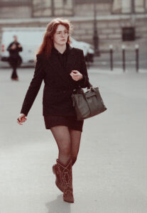 Fannie Girardot, with long hair, walks outdoors in an urban street setting wearing glasses, a black blazer and skirt, patterned cowboy boots, sheer tights, and carrying a dark handbag.