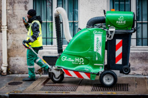 A street cleaner in a yellow safety vest walks beside a green electric street cleaning machine labeled "Glutton H2O" and "Paris Propreté" on a city sidewalk, drinking from a cup.