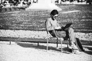 Sebastien Blaise, wearing sunglasses, sits on a chair outdoors working on his laptop, with a backpack beside him. The park or garden setting features people and lush greenery in the background.