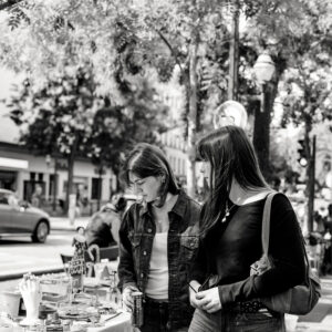 Tiffanie and Pauline stand at an outdoor market table, looking at various small items for sale. Trees and buildings are visible in the background, and a car passes by on the street. The image is in black and white.