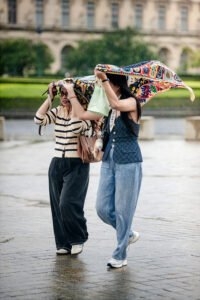 Two women, Dionne and Melissa, walk outside on a rainy day, shielding their heads with a colorful scarf. One wears a striped sweater and glasses; the other opts for a vest and loose jeans. Wet pavement and lush greenery frame the scene.