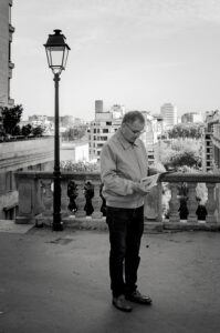 A man wearing glasses and a jacket stands on a city terrace, reading a book—perhaps by Xavier Kieffer. A streetlamp and railing are beside him, with tall buildings and trees in the background under a partly cloudy sky. The image is in black and white.