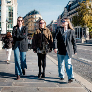 Enya, Camille, and Sabrina walk confidently on a city street lined with historic buildings under a blue sky. Wearing sunglasses and fashionable jackets, one holds a phone; all appear stylish and relaxed.