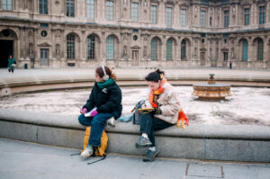 Emmanuelle and Anelise sit on the edge of a fountain outside a historic building. One looks to the side wearing headphones, while the other reads a book. Both have backpacks and wear jackets and scarves.