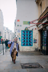 Himari Takahashi, in a coat and scarf, walks on a city sidewalk beside a graffiti-covered wall with a large colorful geometric mural. Other pedestrians and buildings are visible in the background.