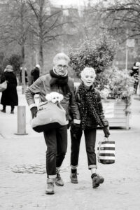 An older couple, Antoine & Celine, stroll outdoors in glasses, jackets, and scarves. The man carries a small white dog in a bag while the woman holds a striped shopping bag. Leafless trees and people fill the background.