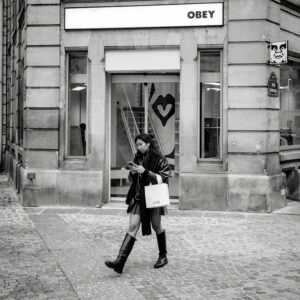 A woman in a dark coat and boots walks past a store with an "OBEY" sign, carrying a shopping bag and looking at her phone; cobblestone street and stone building in the background.
