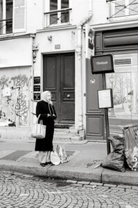 A woman in a headscarf and long coat stands on a cobblestone city sidewalk near a bus stop, reminiscent of Yvonne Derrien's urban photography. Behind her, buildings covered in graffiti and posters complete the black-and-white scene.