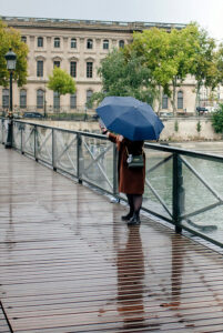 A person in a brown coat holding a blue umbrella stands on a wet wooden bridge, taking a selfie. Across the river, trees and an old building add charm to the scene—just as Gabriel Daniel might have captured in his travels.