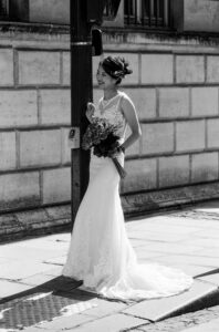Sophia-Marie Lee, in a lace wedding dress, stands on a sunlit city sidewalk holding a bouquet and smiling, with a stone building in the background.