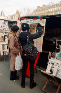 Two women browse through art prints at an outdoor market stand. One points to a large cat print held by Leonie Le Galle, while the other holds a shopping bag. Artworks and posters are displayed around them.