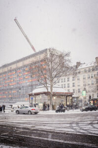 A snowy city street scene in neige Paris, janvier 2026, shows people walking, cars driving, and a motorbike parked. A large building with orange window panels and a crane rises in the background as snow blankets rooftops and bare trees.