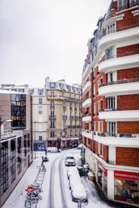 A snowy city street curves between classic Parisian-style apartment buildings in janvier 2026, with parked cars and bicycles covered in neige Paris, and a shop on the corner; the sky is overcast.