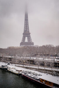 The Eiffel Tower rises into a foggy sky above snow-covered trees and boats along the Seine River in Paris, capturing the magic of neige Paris in janvier 2026.