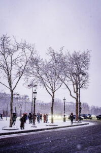 People walk and gather in a snowy park in Paris, lined with bare trees under a cloudy, purple-tinged sky. Neige blankets the ground as street lamps line the path, with buildings visible in the background—capturing Janvier 2026.
