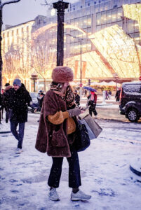 A woman in a brown fur hat and coat stands on a snowy city street in janvier 2026, checking her phone. She holds shopping bags as people walk by. Neige Paris glistens under festive lights illuminating the buildings behind her.