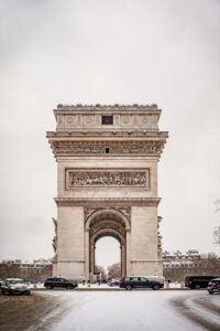 The Arc de Triomphe in Paris stands tall under an overcast sky, as neige recovers the streets in janvier 2026, with several cars parked in the snowy foreground and buildings visible in the background.