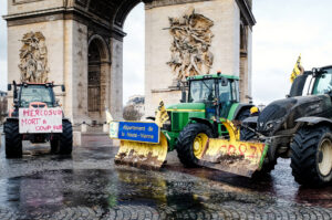 Three tractors driven by agriculteurs with protest signs block a wet cobblestone street near the Arc de Triomphe during a manifestation. The signs, written in French, stand out against the rainy Parisian backdrop.