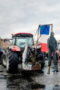 During a manifestation by agriculteurs on a wet, overcast day, a red tractor displays a painted “BREXIT” sign, a mannequin in overalls hangs from a frame, and a blue and red flag with a white sickle symbol waves near the Arc de Triomphe.