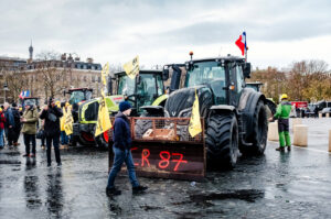 A row of tractors with protest signs and flags, including a French flag, is parked on a city street near the Arc de Triomphe. Agriculteurs in yellow vests stand nearby, holding banners under a cloudy sky.