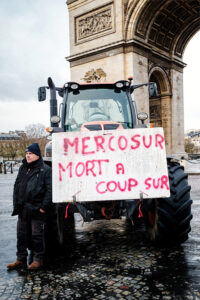 A man stands beside a tractor in front of the Arc de Triomphe during a manifestation by agriculteurs. The tractor has a sign reading "MERCOSUR MORT À COUP SÛR" in red paint, highlighting their protest on a cloudy Paris day.