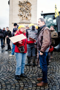 A group of protesters stands on a cobblestone street during a manifestation; a woman in a red jacket and yellow hat holds a cardboard sign while talking to a man in a brown jacket. Other people, agriculteurs, and a tractor are visible in the background.