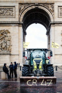 During a manifestation by agriculteurs, a tractor with yellow flags is parked in front of the Arc de Triomphe in Paris, with three people beside it and "CR 47" spray-painted on its front blade.