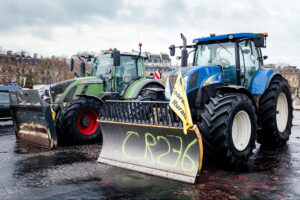 Two large tractors with snowplow blades are parked side by side on wet cobblestone, one bearing “CR376” and a protest sign in French from les agriculteurs. Urban buildings and the Arc de Triomphe can be seen in the background.