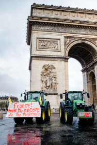 Two green tractors, part of a manifestation by agriculteurs, are parked in front of the Arc de Triomphe. A handwritten banner on one tractor reads, "France! Veux tu encore de tes paysans?" in red and blue letters.