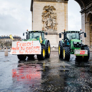 Two green tractors, symbols of the agriculteurs’ manifestation, are parked before the Arc de Triomphe in Paris. One displays a banner: "France! Veux-tu encore de tes paysans?" The wet ground beautifully reflects the iconic scene.