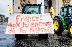 A large white banner with red spray-painted French text is attached to a green tractor in front of the Arc de Triomphe during a manifestation. The sign reads: "France! Veux-tu encore de tes paysans?" Yellow flags are attached to the tractors.