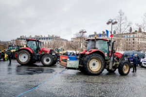 Two red tractors are parked on a wet city square during a manifestation by agriculteurs, surrounded by people and some police. French flags and older buildings are visible in the background under a cloudy sky.