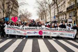 A large group marches down a city street during a manifestation Paris, holding a banner reading "Maltraiter les soignants, c'est maltraiter les patients!" Many soignants libéraux display signs and props, including a pink broken heart.