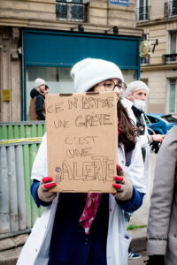 At a manifestation Paris, a person in a white coat, hat, and red gloves—likely one of the soignants libéraux—holds a cardboard sign in French reading, “Ce n’est pas une grève, c’est une alerte,” during a street protest in the city.