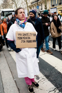 A woman in a white coat joins a manifestation Paris, holding a cardboard sign in French that reads: "Ma santé: j'y tiens, mon médecin: j'en prends soin," with soignants libéraux and others walking behind her.