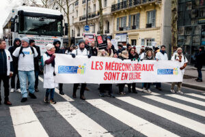 During a manifestation paris, soignants libéraux march on a city street holding a banner that reads “LES MÉDECINS LIBÉRAUX SONT EN COLÈRE.” Some participants, including médecins libéraux, are dressed in white lab coats.