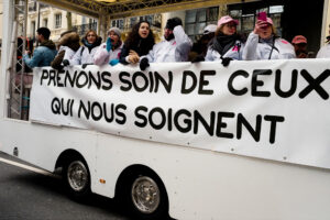 A group of soignants libéraux in white coats stand on a parade float holding a banner that reads, "PRENONS SOIN DE CEUX QUI NOUS SOIGNENT" during a manifestation paris on a city street.