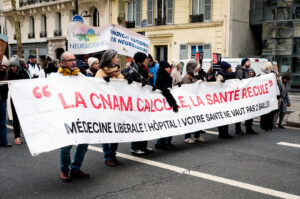A group of people march on a city street during a manifestation Paris, holding a large banner in French that criticizes healthcare cuts, with signs advocating for soignants libéraux, neurologists, and public hospitals visible in the background.