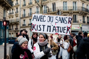 A group of protesters, including soignants libéraux in white coats, stand on a city street during a manifestation Paris, holding a French sign with Parisian buildings in the background.