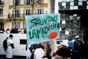 A protester holds a handmade sign reading "SAUVONS LA MEDECINE" (Save Medicine) in French as soignants libéraux in white coats and winter clothing gather on a Paris city street with buildings in the background.