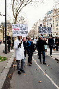 A group of people, including soignants libéraux, march down a city street during a manifestation Paris, holding protest signs in French. The front sign says, "Bonne nouvelle: crever va bientôt coûter moins cher." It's cloudy with bare trees and buildings in the background.