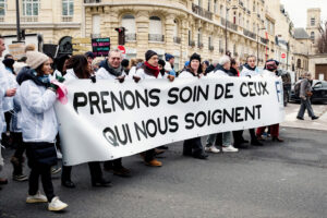 A group of people in white coats, including soignants libéraux, hold a large banner reading "PRENONS SOIN DE CEUX QUI NOUS SOIGNENT" during a manifestation paris with historic buildings in the background.