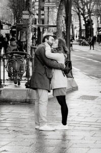 Noel and Samson embrace and kiss on a city sidewalk in winter clothing. People walk by and trees line the street in the busy urban background. The image is in black and white.