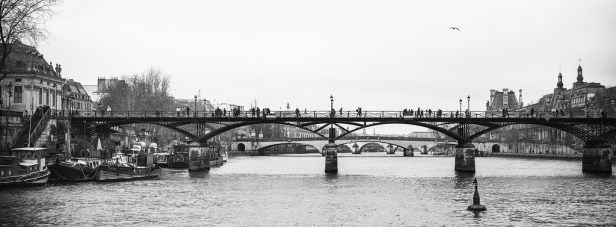 Photo en noir et blanc du pont des Arts sur la Seine à Paris, avec des promeneurs et des bateaux amarrés le long de la rive gauche ; des ponts et des bâtiments historiques lointains apparaissent à l'arrière-plan.