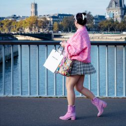 A woman in a pink jumper, checked skirt, and lavender boots walks along a riverside bridge carrying shopping bags. Parisian buildings and trees are visible in the background.