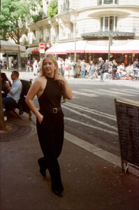 A woman in a black sleeveless jumpsuit walks along a city street with her hands in her pockets. People are gathered on the opposite pavement, and a café with outdoor seating is visible on the left.