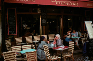 Two elderly women sit and talk at a table outside a Parisian café with woven chairs, under a red canopy. Other customers are seated in the background, and chalkboard menus are visible by the entrance.