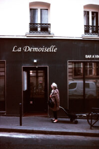 Une femme âgée aux cheveux courts se tient sur un trottoir, tirant un petit caddie, devant une devanture noire sur laquelle est écrit "La Demoiselle". Le reflet d'une voiture est visible dans la vitrine.