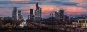 A view of a modern city skyline at dusk, featuring tall glass skyscrapers and office blocks, with a motorway running through the centre and dramatic pink and purple clouds in the sky.